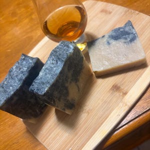 May include: Three handmade soaps with a marbled grey and white design are displayed on a wooden cutting board, alongside a glass of amber-coloured liquid. The soaps have a rustic, natural appearance. The glass is tilted, suggesting a drink.