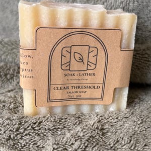 Puede incluir: Una pastilla rectangular de jabón de sebo con una etiqueta de papel marrón. La etiqueta dice "SOAK + LATHER" y "CLEAR THRESHOLD TALLOW SOAP Net. 5oz.". El jabón es de color amarillo claro y tiene una parte superior texturizada.