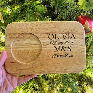 Könnte beinhalten: Rechteckiges Holzservierbrett mit runder Vertiefung. Auf dem Brett steht der Text "OLIVIA Off my tits on M&S Picky Bits". Das Brett wird vor einem Weihnachtsbaum gehalten.