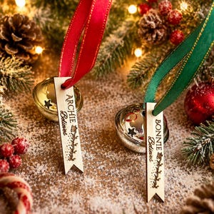 May include: Two silver-toned jingle bells attached to light-colored wooden tags with red and green ribbons. The tags are engraved with the names Archie and Bonnie, the word Believes, and a small reindeer graphic with a trail of stars.