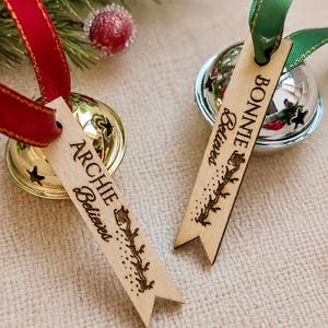 May include: Two metal jingle bells with attached wooden tags. One gold bell has a red ribbon and a tag reading ARCHIE Believes with a sleigh silhouette. One silver bell has a green ribbon and a tag reading BONNIE Believes with a sleigh silhouette.