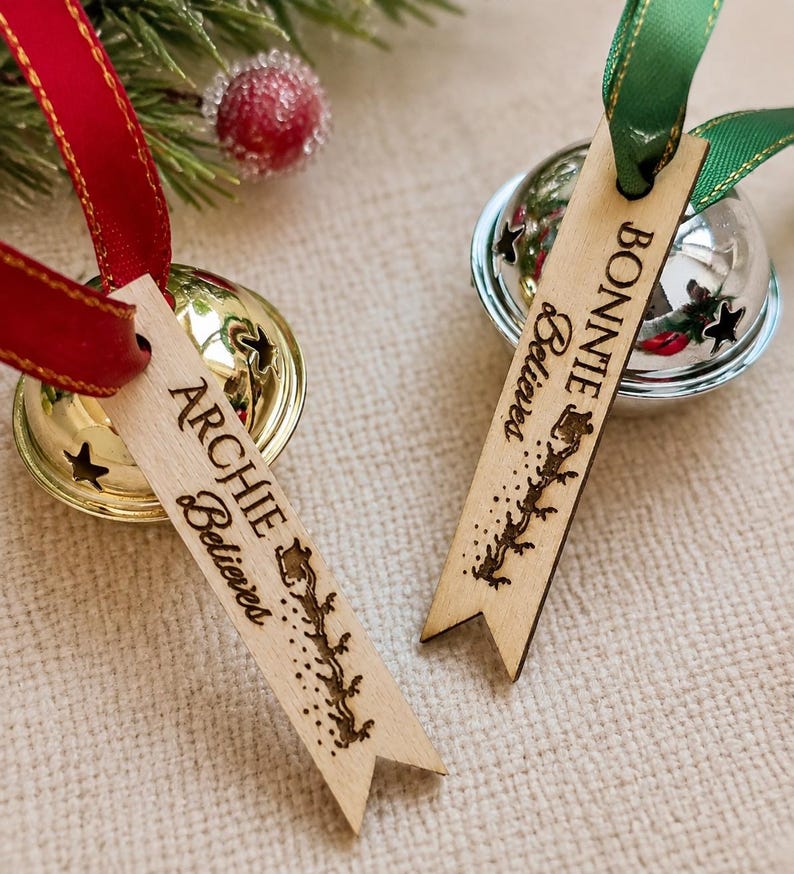 May include: Two metal jingle bells with attached wooden tags. One gold bell has a red ribbon and a tag reading ARCHIE Believes with a sleigh silhouette. One silver bell has a green ribbon and a tag reading BONNIE Believes with a sleigh silhouette.