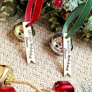 May include: Close-up of Christmas bells with red and green ribbons. One bell has a tag that reads "Archie Believes" and the other reads "Bonnie Believes". The bells are gold and silver, with a star design.