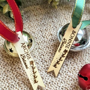May include: Two silver jingle bells with attached wooden tags. One tag features the name Archie and the text Believes with a reindeer silhouette. The other tag features the name Bonnie and the text Believes with a reindeer silhouette. Red and green ribbons are attached to the bells.