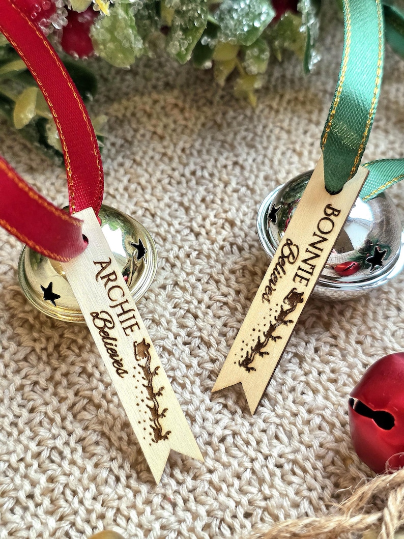 May include: Two silver jingle bells with attached wooden tags. One tag features the name Archie and the text Believes with a reindeer silhouette. The other tag features the name Bonnie and the text Believes with a reindeer silhouette. Red and green ribbons are attached to the bells.