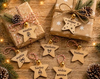 May include: Six light-colored wooden star ornaments featuring engraved names and holiday icons, including a snowman, reindeer, bow, Santa, and sleigh. Each star has a gold bell and red and white twine, with two stars attached to brown gift boxes.