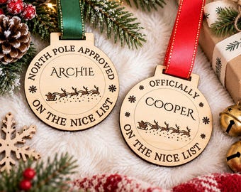 May include: Two circular wooden ornaments featuring engraved text and graphics. One ornament says North Pole Approved Archie On The Nice List with a green ribbon. The other says Officially Cooper On The Nice List with a red ribbon.