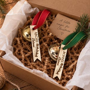 May include: Two gold jingle bells with star cutouts, each attached to a wooden tag engraved with the names Archie and Bonnie and the word Believed. The tags feature red and green ribbons and are presented in a box with a card reading A little magic for Christmas.
