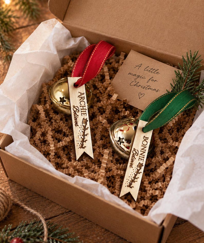 May include: Two gold jingle bells with star cutouts, each attached to a wooden tag engraved with the names Archie and Bonnie and the word Believed. The tags feature red and green ribbons and are presented in a box with a card reading A little magic for Christmas.