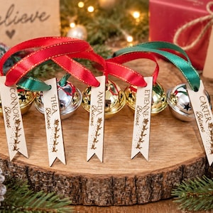 May include: Five metallic jingle bells in gold and silver finishes, each attached to a red or green ribbon with a personalized wooden tag. The tags are engraved with names including Connor, Thea, Amelia, Etienne, and Carter, plus the word Believes.
