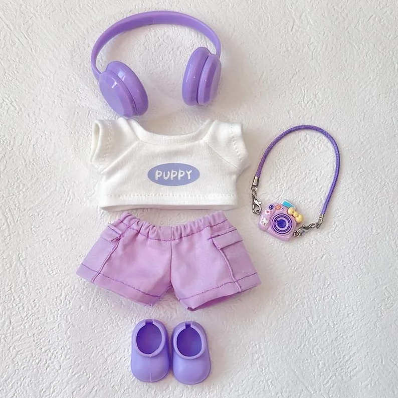 Op de afbeelding: Een poppenoutfitset in paars en wit. De set bevat een wit t-shirt met het woord "PUPPY", een paarse korte broek, schoenen, een koptelefoon en een cameravormige tas. De items zijn op een witte ondergrond geplaatst.