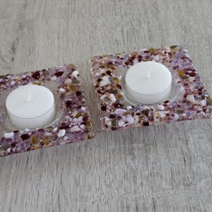 Puede incluir: Dos candelabros cuadrados con un diseño de mosaico de piedras moradas, blancas y marrones. Cada soporte tiene una vela de té blanca en el centro. Los candelabros están sobre una superficie de madera gris claro.