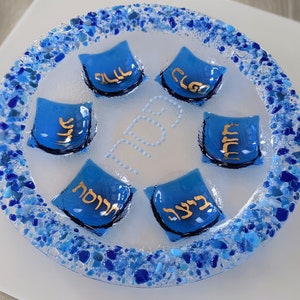 May include: A blue glass Seder plate with a white background and a blue and white mosaic border. The plate has five sections, each with a blue pillow-shaped piece of glass with Hebrew writing in gold.