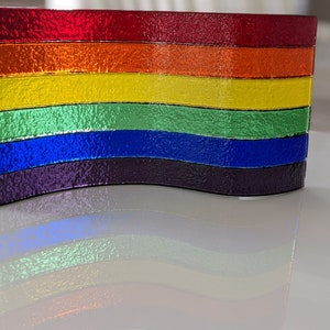 May include: A rainbow-colored glass art piece with a wavy design. The piece is made of six horizontal stripes of glass in the colors red, orange, yellow, green, blue, and purple.