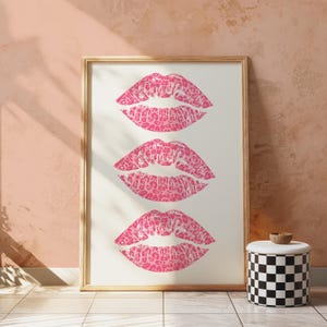 Framed wall art print featuring three pink lips with a leopard print design, arranged vertically on a white background, leaning against a pink paneled wall in a well-lit room.