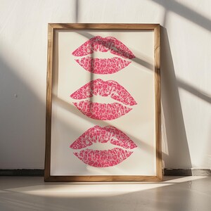 Framed wall art print featuring three pink lips with a leopard print design, arranged vertically on a white background, leaning against a pink paneled wall in a well-lit room.