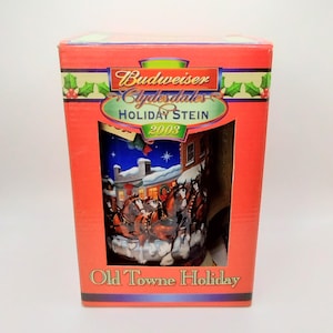 Può includere: Una scatola rossa con un bordo bianco e un'immagine di una boccale di birra Budweiser Clydesdales Holiday Stein. Il boccale presenta una scena invernale con i Clydesdales che tirano un carro. La scatola dice "Budweiser Clydesdales Holiday Stein 2003" e "Old Towne Holiday".
