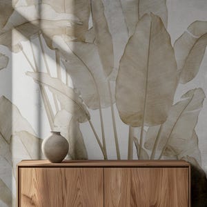 May include: A wooden cabinet with two doors sits in front of a wall with a large-scale botanical print featuring beige banana leaves. The cabinet has a light brown wood grain finish.
