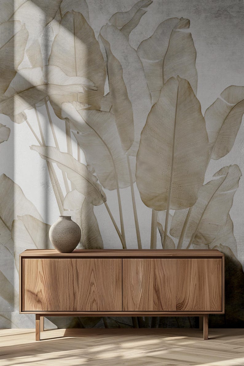 May include: A wooden cabinet with two doors sits in front of a wall with a large-scale botanical print featuring beige banana leaves. The cabinet has a light brown wood grain finish.