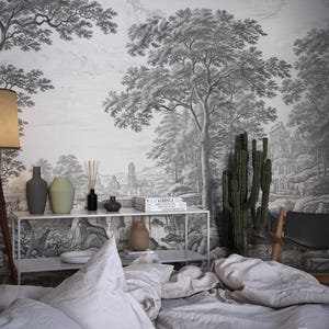 May include: A bedroom interior with a black and white wallpaper depicting a forest scene. The wallpaper features trees, a path, and a building in the distance. A bed with white linens is in the foreground, and a cactus plant is in a pot on the floor. There is a table with a lamp, vases, and books on it.