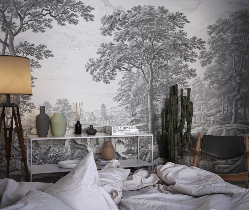 May include: A bedroom interior with a black and white wallpaper depicting a forest scene. The wallpaper features trees, a path, and a building in the distance. A bed with white linens is in the foreground, and a cactus plant is in a pot on the floor. There is a table with a lamp, vases, and books on it.