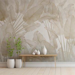 May include: A light beige wall with a large-scale botanical print of leaves in a muted beige tone. A wooden console table with a white ceramic vase and two smaller white ceramic vases sits in front of the wall. Two potted green plants are on the floor in front of the table.