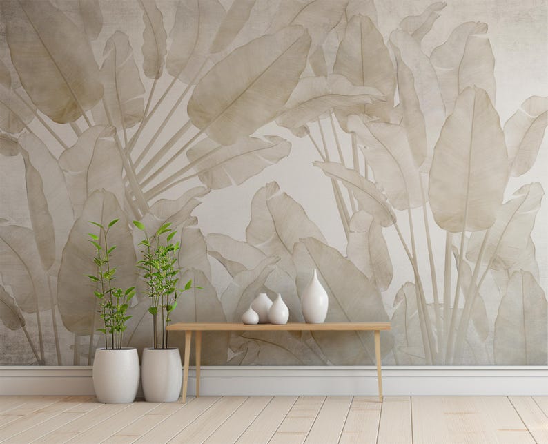 May include: A light beige wall with a large-scale botanical print of leaves in a muted beige tone. A wooden console table with a white ceramic vase and two smaller white ceramic vases sits in front of the wall. Two potted green plants are on the floor in front of the table.