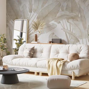May include: A white sofa with a light brown throw blanket and a tan pillow sits in a living room with a large wall mural of beige leaves. A black coffee table with a magazine and glasses sits in front of the sofa.