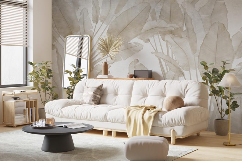 May include: A white sofa with a light brown throw blanket and a tan pillow sits in a living room with a large wall mural of beige leaves. A black coffee table with a magazine and glasses sits in front of the sofa.