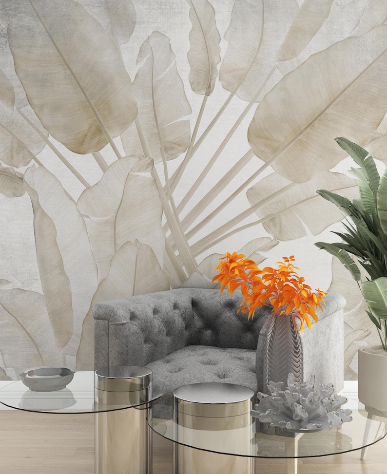 May include: A gray tufted armchair with a glass coffee table and two smaller side tables. The room has a white wall with a large print of beige banana leaves. A vase with orange flowers sits on the table.
