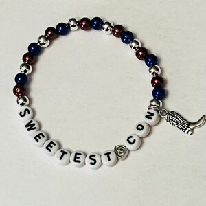 May include: A beaded bracelet with red, white, and blue beads and a charm of a boot. The bracelet spells out "Sweetest Con".