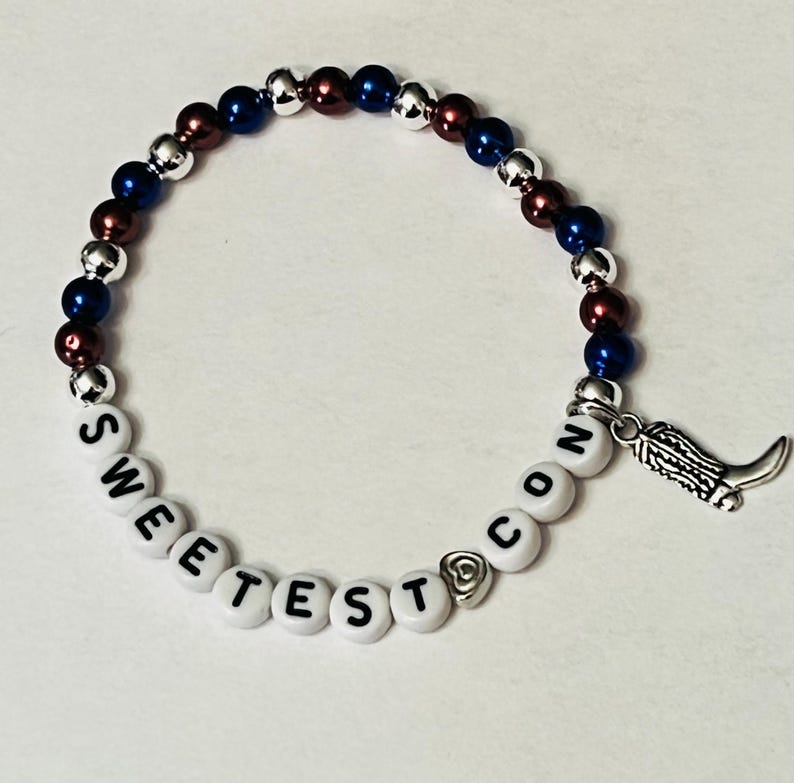 May include: A beaded bracelet with red, white, and blue beads and a charm of a boot. The bracelet spells out "Sweetest Con".