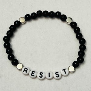 May include: Black beaded bracelet with silver beads and white letter beads spelling out the word "RESIST".
