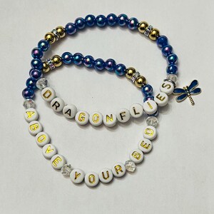 May include: Two beaded bracelets, one blue with gold accents and a dragonfly charm, the other white with gold lettering spelling "ABOVE YOUR BED" and "DRAGONFLIES".