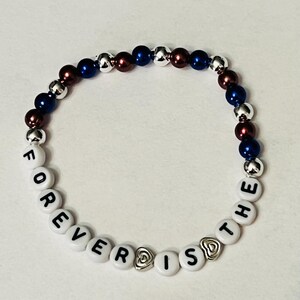 May include: A white beaded bracelet with the words "FOREVER IS THE" spelled out in black letters. The bracelet is accented with silver and blue beads, and two silver heart charms.