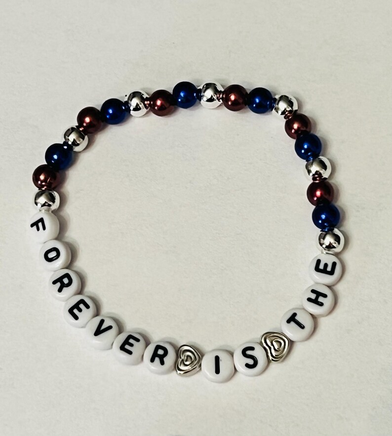 May include: A white beaded bracelet with the words "FOREVER IS THE" spelled out in black letters. The bracelet is accented with silver and blue beads, and two silver heart charms.