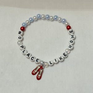 May include: A beaded bracelet with the phrase "NO PLACE LIKE HOME" spelled out in white letter beads. The bracelet features red and blue beads, and a charm of red ruby slippers.
