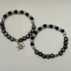 May include: Two black and silver beaded bracelets. One bracelet spells "LIGHTS CAMERA" and has a silver camera charm. The other bracelet spells "BITCH SMILE".