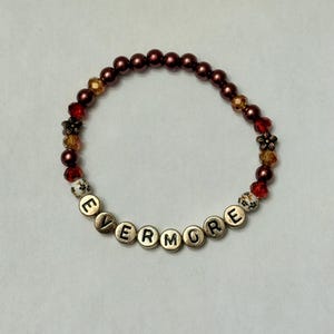 May include: A beaded bracelet with the word "EVERMORE" spelled out in gold letter beads. The bracelet features maroon, red, and gold beads, along with decorative flower-shaped beads. The bracelet is designed to be worn on the wrist.