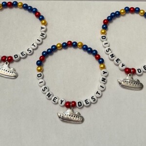 May include: Three beaded bracelets featuring alternating red, blue, and yellow spherical beads. Each bracelet includes white letter beads spelling out DISNEY DESTINY and a silver-toned metal charm shaped like a cruise ship hanging from the center.