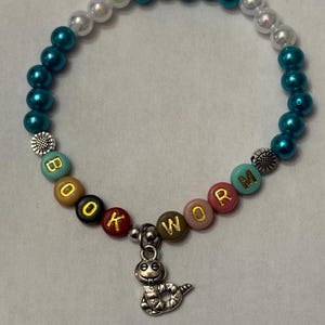 May include: A beaded bracelet with teal, white, and colorful beads spelling "BOOK WORM." The bracelet features a silver charm of a smiling worm. The beads are strung on an elastic band.