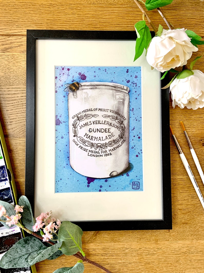 Framed watercolour artwork of a jar of Keillers Dundee marmalade with a bee on a blue and purple splattered background, on a wooden table surrounded by paintbrushes and flowers