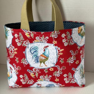 May include: A fabric tote bag with a red background featuring a rooster design. The bag has a floral pattern with white accents and a blue interior. It has olive green handles.