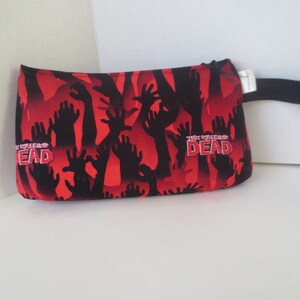 May include: Red zippered pouch with a black strap. The pouch has a repeating pattern of black zombie hands reaching up against a red background. The text "The Walking Dead" is printed on the pouch.