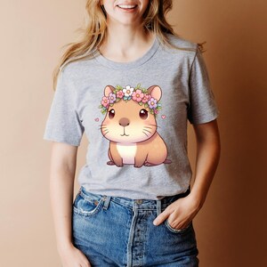 A lady in a grey tshirt featuring a digital download image of a kawaii capybara that has a tiny heart on each side of the outside of the image And a capybara has a floral headband around its head with pink purple and white flowers.