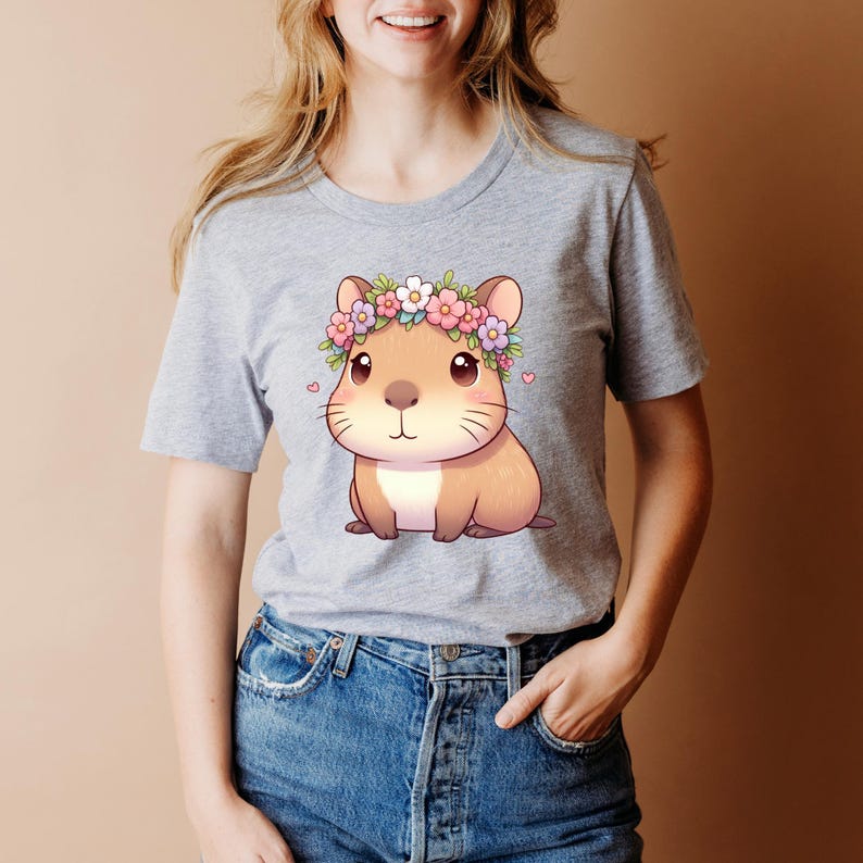 A lady in a grey tshirt featuring a digital download image of a kawaii capybara that has a tiny heart on each side of the outside of the image And a capybara has a floral headband around its head with pink purple and white flowers.