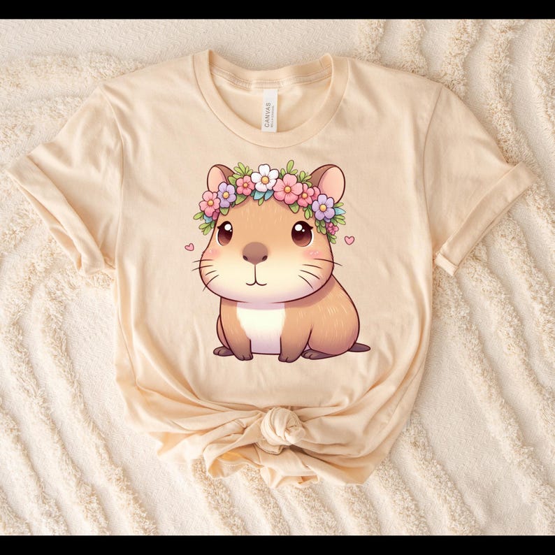A beige T-shirt with a digital download image of a capybara that has a tiny heart on each side of the outside of the image And a capybara has a floral headband around its head with pink purple and white flowers and the shirt is on a beige blanket.
