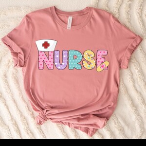 Może przedstawiać: Różowy T-shirt z napisem "NURSE" w kolorowych literach. Litery są ozdobione sercami, symbolami medycznymi i pigułkami. Nad słowem znajduje się czapka pielęgniarki z czerwonym krzyżem.