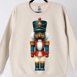 May include: A beige sweatshirt featuring a colorful illustration of a nutcracker soldier. The nutcracker is depicted with a dark blue hat, a gold star, and a red and blue uniform. The design is centered on the front of the sweatshirt.