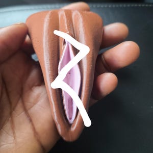 Könnte beinhalten: Eine braune, 3D-gedruckte Skulptur mit rosa Innenraum. Auf der Vorderseite ist ein weißes, eckiges Design gezeichnet. Die Skulptur wird in einer Hand gehalten.